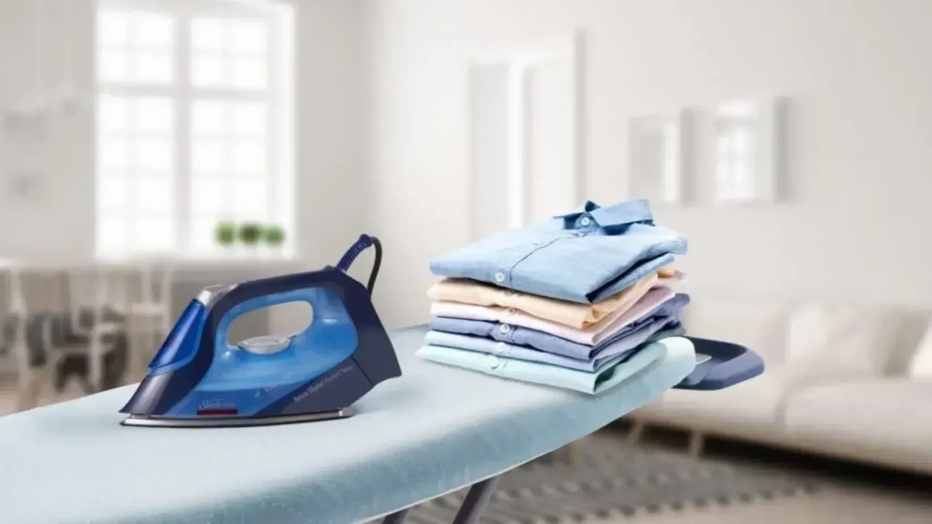 An iron on an ironing board with a pile of freshly pressed shirts