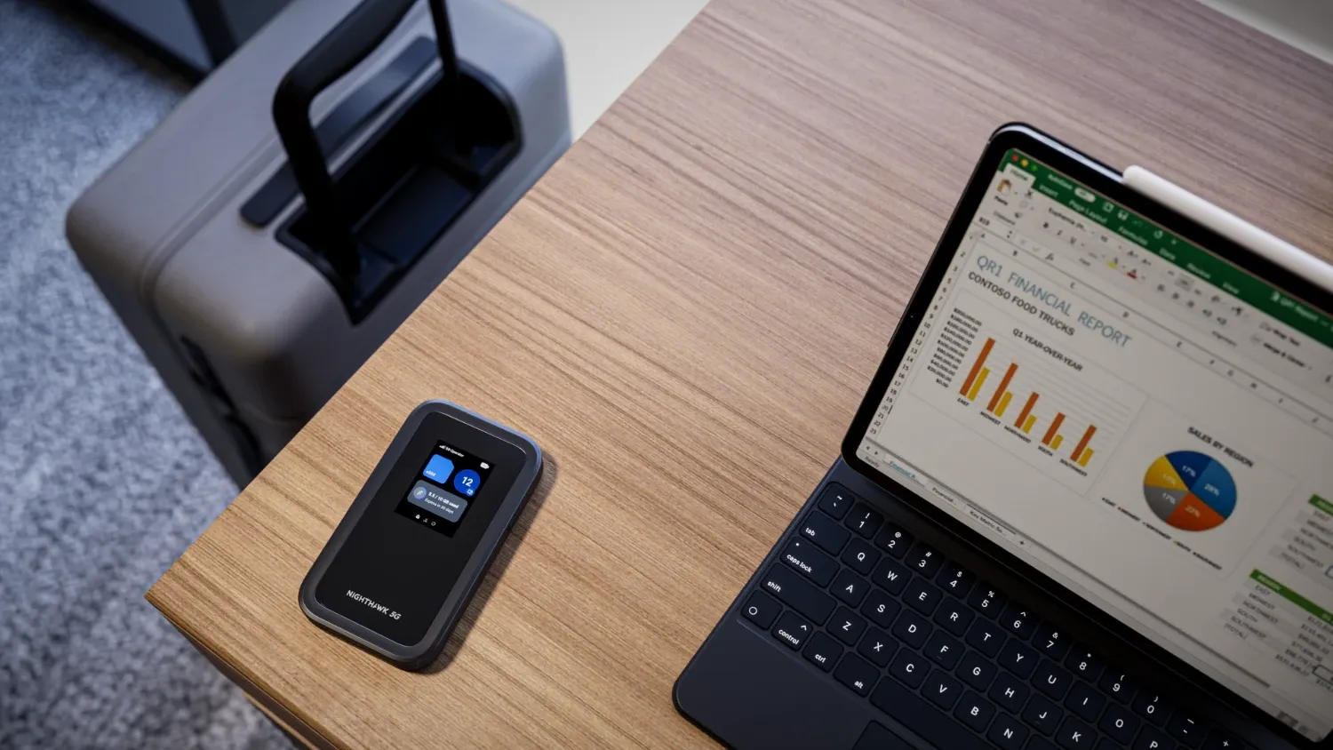 The new Netgear 5G M7 hotspot on a desk next to a laptop with a suitcase on the floor next to the desk