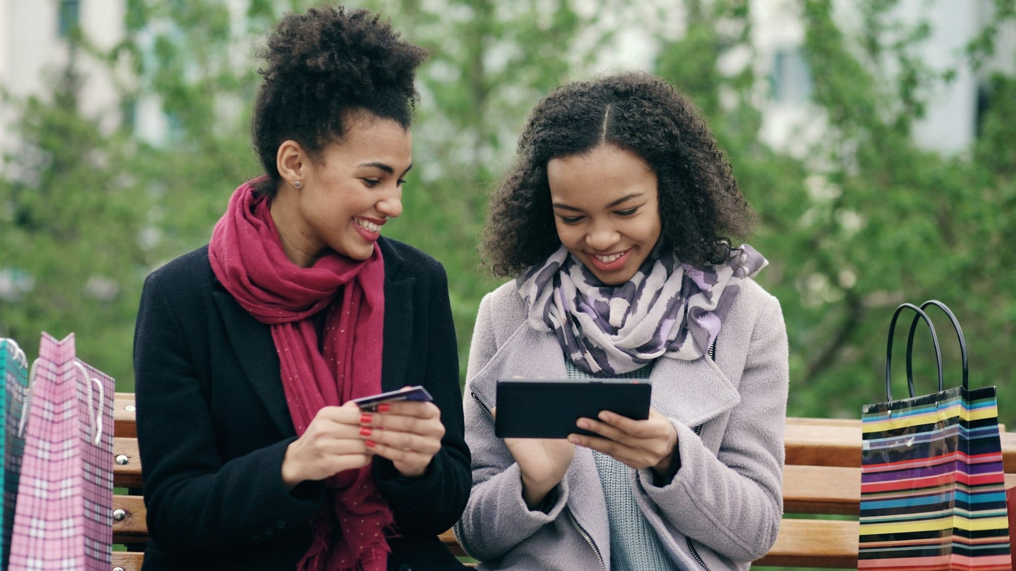 Two women on a park bench, onw with a phone and the other with a credit card. Shopping bags sit on either side.